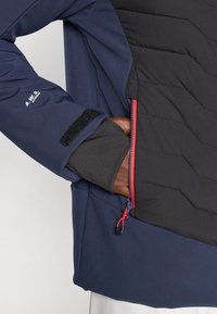 Navy Softshell-Jacke mit schwarzem isoliertem Vorderteil, Reißverschlusstasche mit roten Akzenten und Klettverschluss an den Bündchen. Strukturierter, gesteppter Design.