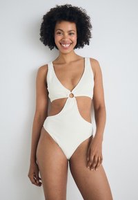 Maillot de bain une pièce blanc à côtes avec décolleté en V profond, côtés découpés et quincaillerie circulaire décorative au centre. Texture douce, design ajusté.