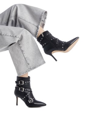 Botas negras hasta el tobillo con punta afilada, hebillas, tachuelas y una cremallera trasera. Tienen un tacón de aguja y se combinan con unos jeans grises de pierna ancha.