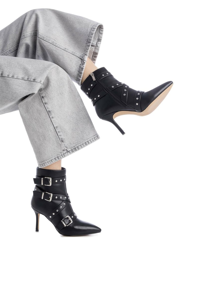 Botas negras hasta el tobillo con punta afilada, hebillas, tachuelas y una cremallera trasera. Tienen un tacón de aguja y se combinan con unos jeans grises de pierna ancha.