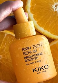 Gul glasflaska med serum och pipettlock, med mörk text som lyder "Skin Tech Serum Brightening Booster", placerad mot bakgrund av apelsinskivor.