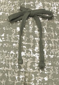 Tecido verde-oliva com padrões de manchas brancas, apresentando um cordão trançado correspondente atado na cintura para ajuste. 