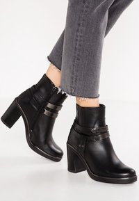 Botas de tobillo de cuero negro con tacón grueso, que cuentan con correas decorativas y una cremallera lateral. Textura suave y diseño estructurado.