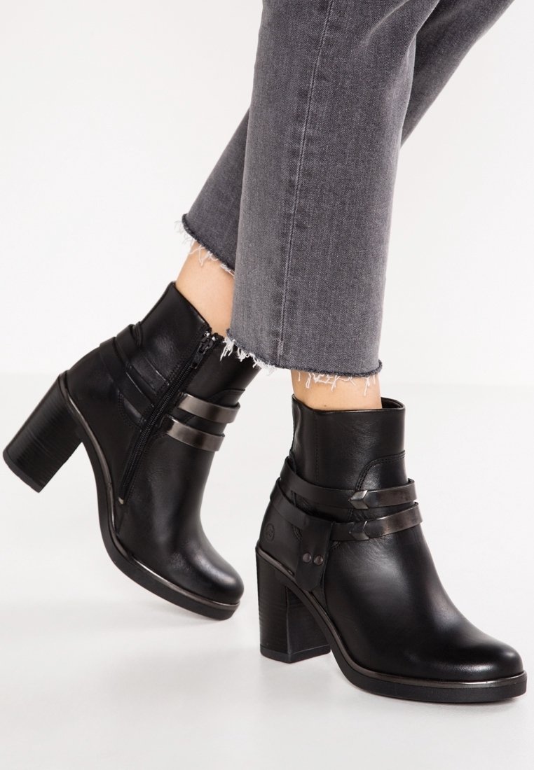 Botas de tobillo de cuero negro con tacón grueso, que cuentan con correas decorativas y una cremallera lateral. Textura suave y diseño estructurado.