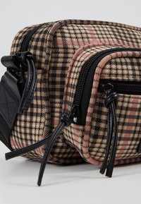 Ruskea ja musta ruudullinen olkalaukku vaaleanpunaisin yksityiskohdin, jossa kaksi vetoketjulla suljettavaa lokeroa sekä mustaa nahkaa olevat yksityiskohdat ja hihnat.