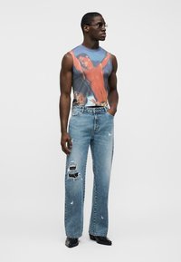 Αμάνικο τοπ με γραφικό σχέδιο σε μπλε φόντο και εκτύπωση στον κορμό. Φορεμένο με φθαρμένο ανοιχτό μπλε τζιν και μαύρα μυτερά παπούτσια.