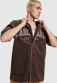Camicia in velluto a coste marrone con maniche corte, caratterizzata da dettagli bianchi, grandi lettere "PARL." e "EST 89", e bottoni neri lungo la parte anteriore.