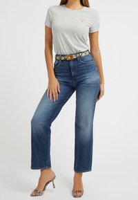 T-shirt grigia con piccolo logo, jeans in denim blu a vita alta e cintura con motivo a serpente. Sandali con tacco trasparente e punta aperta.