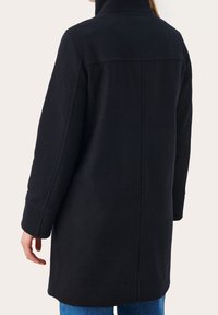 Sort frakke med høj krave, lige snit og lange ærmer. Har rene linjer og et blødt, struktureret stof. Bæres over blå jeans.