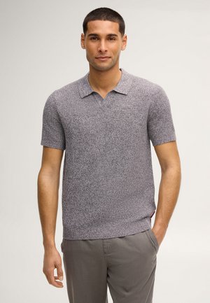 Uomo con capelli corti che indossa una polo grigia a maniche corte con texture e pantaloni grigi, in piedi con una mano in tasca.