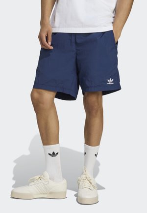 Mężczyzna w granatowych krótkich spodenkach Adidas, białych skarpetach sportowych Adidas oraz beżowych tenisówkach Adidas, stojący z rękami w kieszeniach na białym tle.