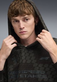 Felpa con cappuccio scura a motivo geometrico, con logo PUMA e scritta "HIGH SPEED, HIGH STAKES" in blu sul petto e sul cappuccio.