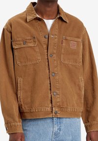 Brązowa dżinsowa kurtka zapinana na guziki z dwoma kieszeniami na piersi, noszona na białej koszuli i jasnoniebieskich dżinsach.