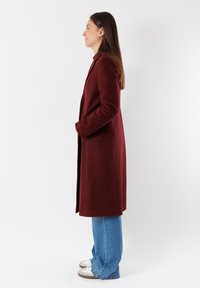 Cappotto di lana bordeaux con maniche lunghe, taglio dritto e tasche laterali, abbinato a jeans blu larghi e sneakers grigie.