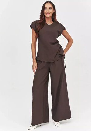 Kobieta stoi z ręką na biodrze, ubrana w ciemnobrązowy marszczony krótki top z krótkim rękawem i szerokie spodnie z białymi spiczastymi butami.