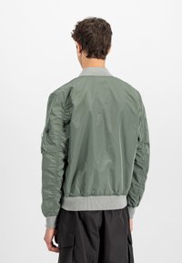 Olive green bomber jacket with a smooth, shiny fabric, ribbed cuffs and collar, utility pocket on the sleeve, and elastic hem.