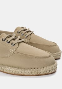 Beige espadrill lábbelik sima velúrból készült felsőrésszel, fűzős dizájnnal és szőtt juta talppal. Kerek orral és sötét fűzőlyukakkal rendelkezik.