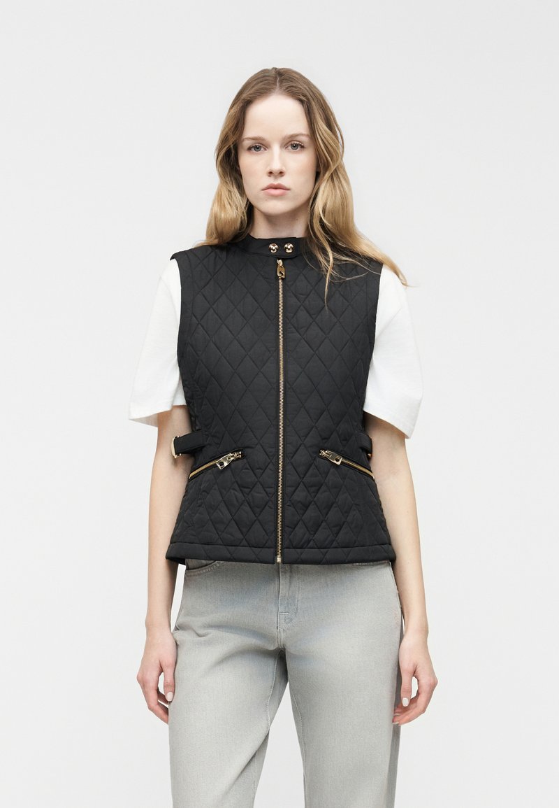 Γυναίκα που φοράει μαύρο αμάνικο γιλέκο με χρυσά φερμουάρ πάνω από λευκό κοντομάνικο πουκάμισο και ανοιχτό γκρι τζιν, στέκεται μπροστά σε λευκό φόντο.