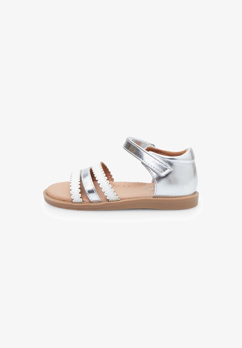 Silver metallic sandal with three straps, decorative scalloped trim, and an adjustable Velcro closure. Flat sole with a brown base.