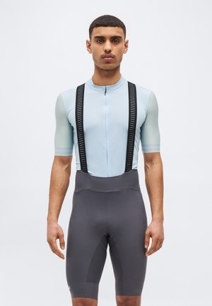 Hombre con camiseta de ciclismo azul claro de manga corta y bib shorts gris oscuro con tirantes negros, de pie contra un fondo blanco.