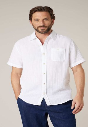 Homme barbu portant une chemise blanche à manches courtes et un pantalon bleu foncé, debout devant un fond clair uni.