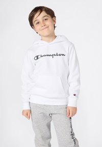 Ung gutt i hvit Champion-hettegenser og grå joggebukser står og smiler svakt mot en enkel lys bakgrunn.