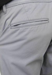 Poche arrière d'un pantalon gris clair avec ouverture passepoilée et coutures visibles sur un tissu lisse.