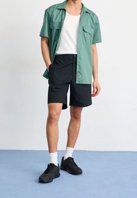 Světle zelená košile s krátkým rukávem a dvěma kapsami, spárovaná s černými šortkami. Model má na sobě černé tenisky a bílé ponožky, stojí na modré podlaze.