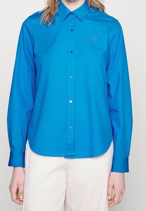 Woman wearing a long-sleeve bright blue button-up shirt with a collar, paired with white pants, shown from neck to hips.