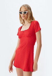 Rode textuur mini-jurk met korte mouwen, vierkante halslijn en een lichte rimpeling bij de buste. G_dragen met witte cat-eye zonnebril.