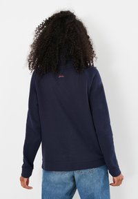 Felpa in pile di colore blu navy con colletto alto, polsini e bordo a costine, caratterizzata da un piccolo logo rosso sul retro. Design a vestibilità casual.