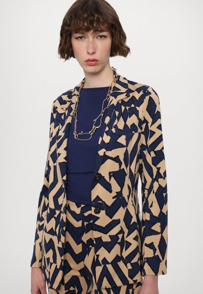 Beige blazer med navy abstrakt mønster, to knapper og klapkraver, parret med en navy top. Har en lagdelt kæde halskæde.