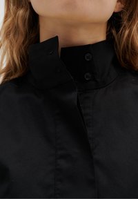 Schwarze Hemd mit Kragen, das ein strukturiertes Design, zwei schwarze Knöpfe und eine glatte Textur aufweist, wobei der Fokus auf dem Hals- und Kragenbereich liegt.