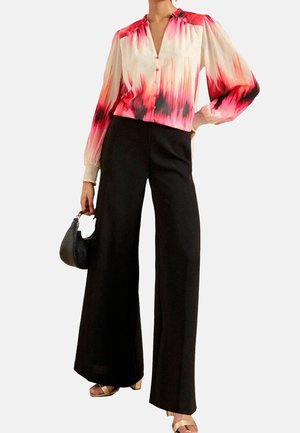 Femme portant un pantalon noir à jambes larges et un chemisier à manches longues avec un dégradé rouge, rose et crème, tenant un petit sac à main noir.
