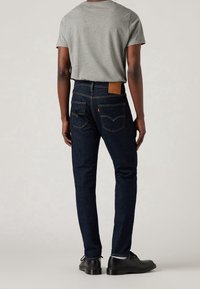 Jeans de mezclilla azul oscuro con costuras amarillas en contraste, cinco bolsillos y parche de cuero en la parte trasera. Combinados con una camiseta de manga corta gris.