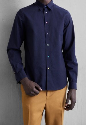 Mężczyzna ubrany w granatową koszulę z długim rękawem z kolorowymi guzikami i musztardowymi spodniami, stojący na tle szarej ściany.