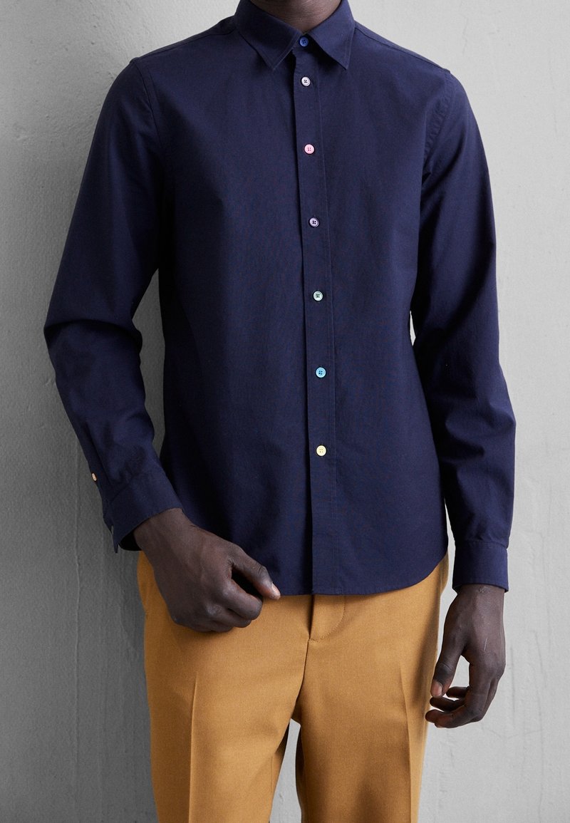 Homme portant une chemise bleu marine à manches longues avec des boutons colorés et un pantalon jaune moutarde, debout devant un mur gris.