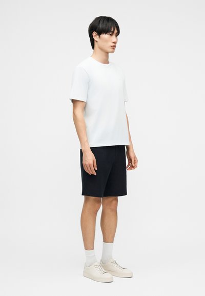 Homme debout portant un t-shirt blanc à manches courtes, un short noir jusqu'aux genoux, des chaussettes blanches et des baskets beiges sur un fond uni.
