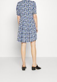 Vestido azul con patrón y mangas cortas, que presenta una mezcla de formas geométricas en negro, blanco y morado. Zapatos de tacón bajo negros.