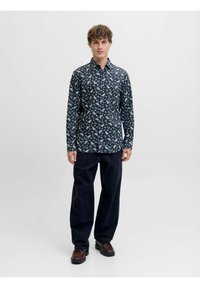 Camicia navy con motivo floreale bianco, colletto a bottone, maniche lunghe e taglio dritto. I pantaloni sono scuri, con vestibilità rilassata. Scarpe marroni.