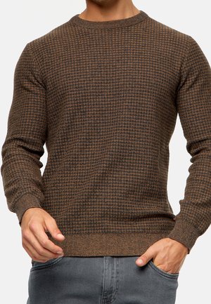Mann trägt braunen, gemusterten Strickpullover mit gerippten Bündchen und Saum, kombiniert mit grauen Jeans, Hände in der Nähe des Bunds.