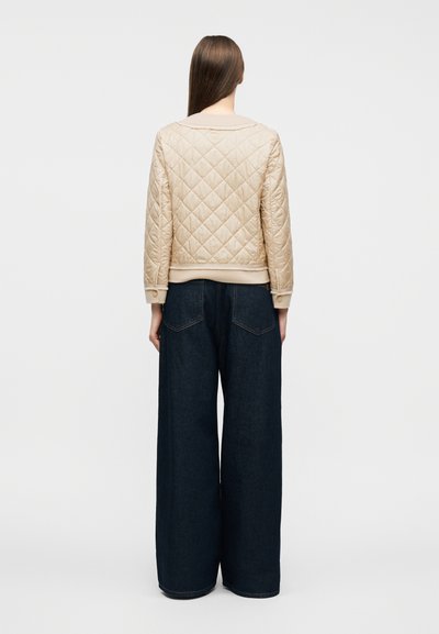 Γυναίκα με μακριά καστανά μαλλιά, φορώντας ένα μπεζ καπιτονέ μπουφάν και σκούρα μπλε τζιν παντελόνι με φαρδιά πόδια, στέκεται πλάτη σε λευκό φόντο.