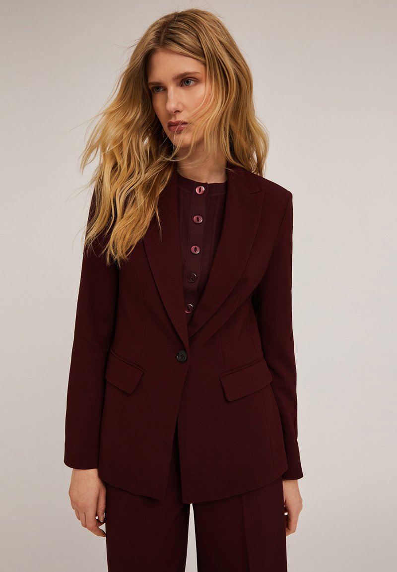 Bordowy dopasowany blezer z subtelnymi klapami, dwoma kieszeniami z przodu, zapięciem na jeden guzik, noszony na dopasowanej bluzce z dużymi guzikami.