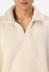 Beige, fluffy fleece pullover med krave og dyb V-hals. Teksturen ser blød og let struktureret ud, hvilket giver et hyggeligt look.