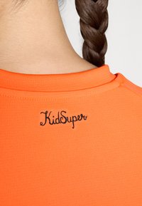 Fényes narancssárga, rövid ujjú póló bordázott textúrával, a hátsó nyakrészen fekete "KidSuper" logóval hímzve.