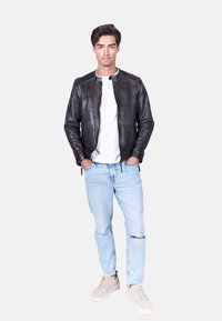 Giubbotto di pelle nera con collo rotondo, tasche laterali con zip e polsini a coste, abbinato a jeans strappati di un blu chiaro e sneakers grigie.