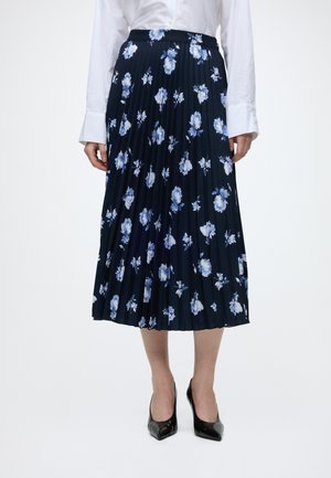Femme portant une jupe midi plissée bleu marine à motif floral bleu clair, une chemise blanche boutonnée et des talons pointus noirs.