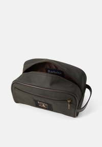 Bolsa de toalete Barbour em verde escuro com bolso frontal com fecho, detalhes em couro e pega de tecido, mostrada parcialmente aberta sobre fundo branco.