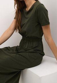 Mörkgrön klänning med korta ärmar, rund hals och elastisk midja. Tillverkad av mjukt tyg, med en flytande siluett och minimal design.