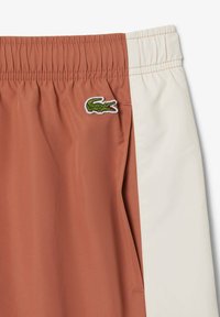 Bañadores marrones y crema con cinturilla elástica, que presentan un logo de cocodrilo bordado en verde en el lado izquierdo y una textura suave.